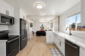 Een moderne keuken met witte kastjes, een zwarte koelkast, een magnetron en een vaatwasser. Er is een zithoek met een tafel en stoelen, en een zwarte bank op de achtergrond.