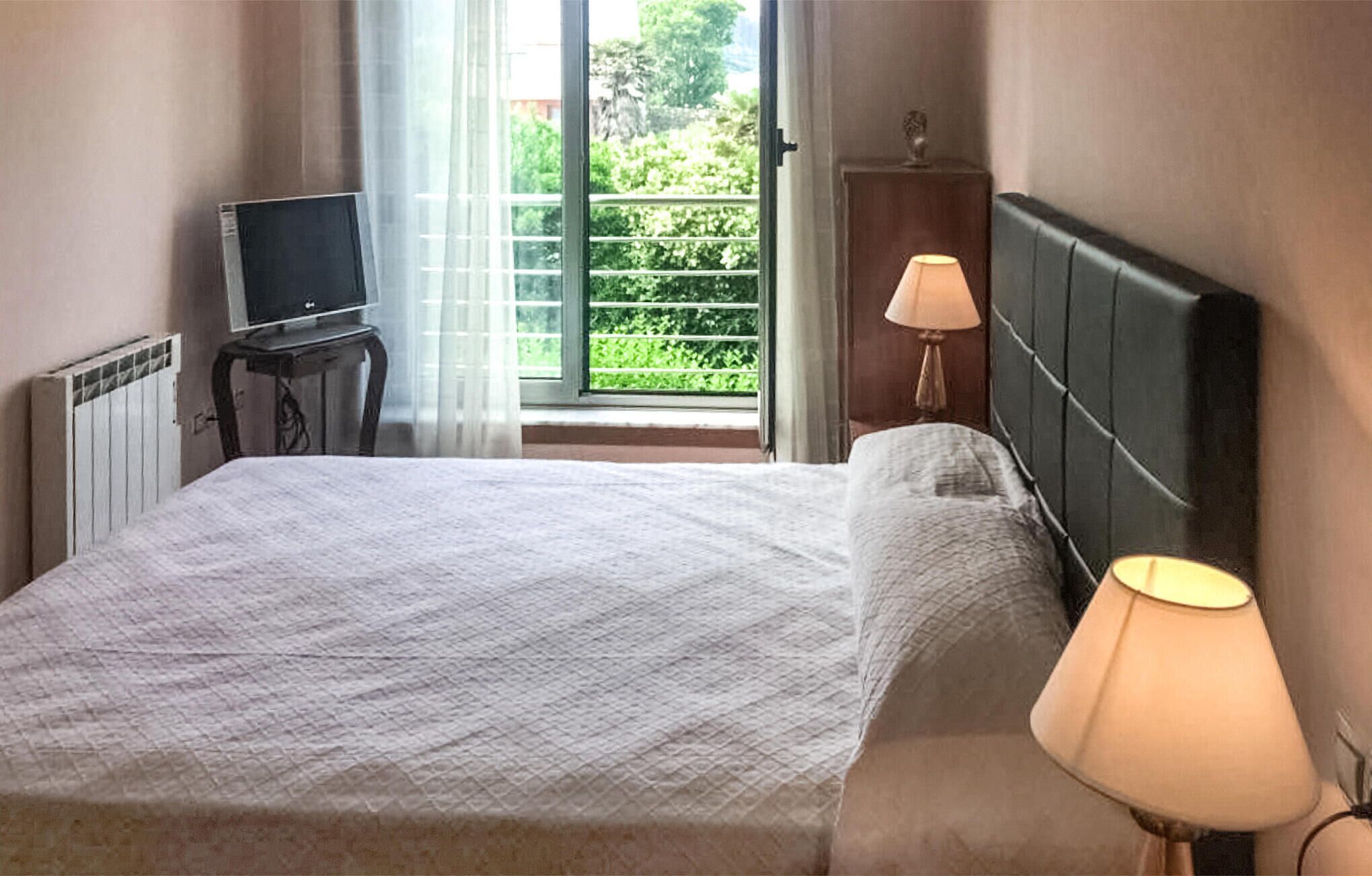 Une chambre d’hôtel avec un lit, un téléviseur posé sur une table de chevet, une lampe et une vue sur la verdure par la fenêtre.