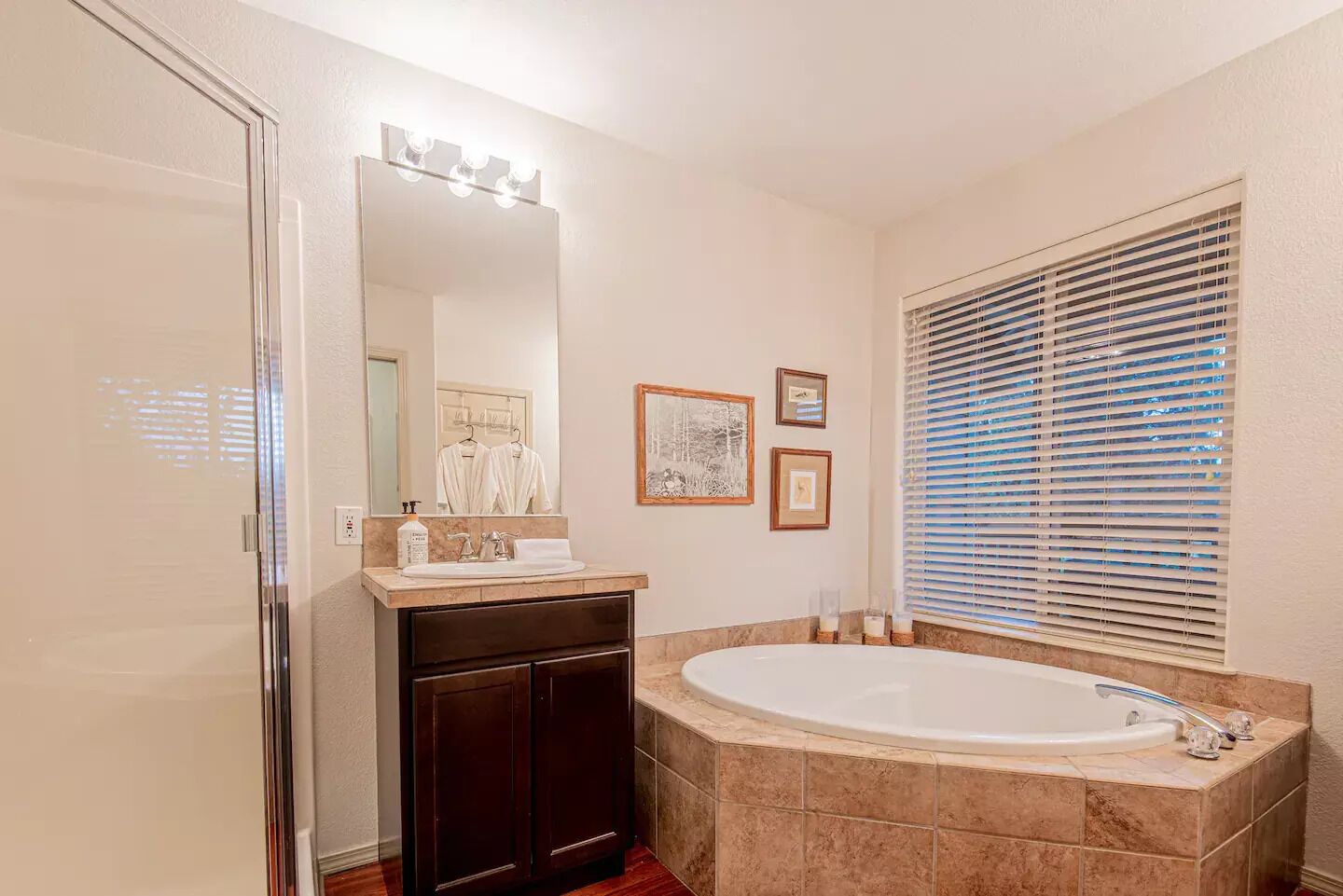 A bathroom with a large bathtub, a vanity with a mirror and lights, and a window with blinds.