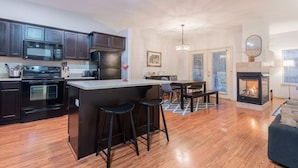 Una cocina moderna con armarios oscuros, una isla central con taburetes, un área de comedor con mesa y sillas, y una chimenea.