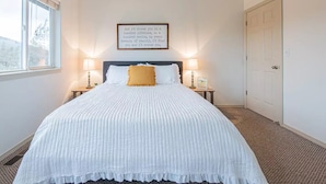 Una cama bien hecha con una colcha blanca y un almohadón amarillo, con dos lámparas de noche a cada lado. Una cita enmarcada cuelga sobre la cama.