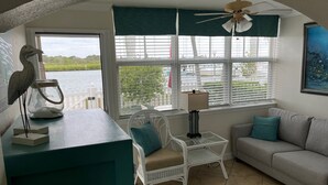 A cozy room with a view of the water, a white chair, a sofa, and a table with a lamp. - May availability: Waterfront corner unit, private patio, first floor (Indian Shores)