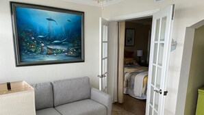 A room with a grey sofa, a painting of an underwater scene, and a bed with a striped blanket. - May availability: Waterfront corner unit, private patio, first floor (Indian Shores)