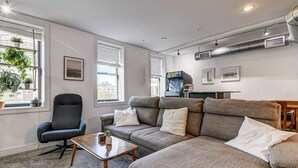 Ein modernes Wohnzimmer mit einem grauen Sofa, einem schwarzen Sessel und einem kleinen Holztisch. Zwei Fenster mit Jalousien, eine Wandklimaanlage und ein gerahmtes Bild sind vorhanden.