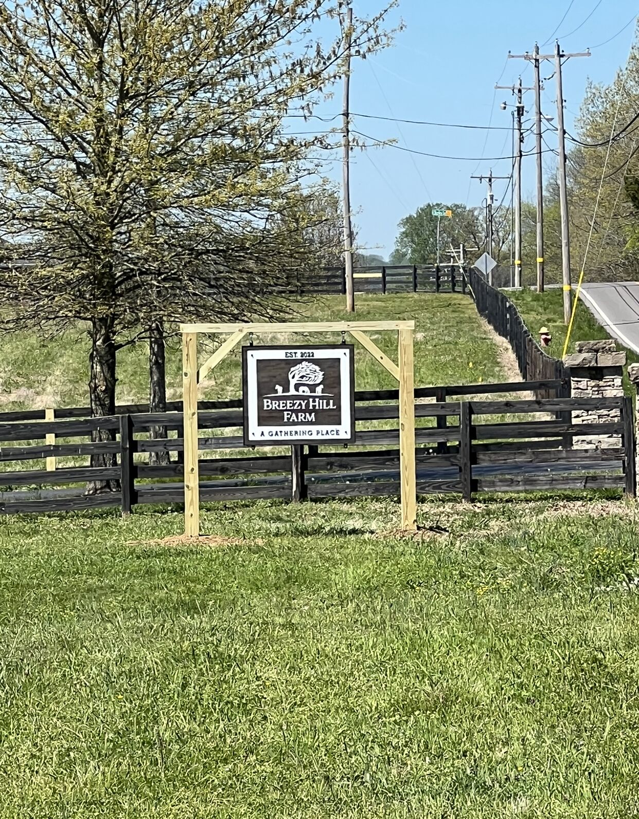 A sign for Breezy Hill Farm, a gathering place, with a wooden fence and a grassy area in the foreground.
