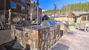 An outdoor grill area with stone walls, surrounded by chairs and tables, and a building in the background. - Grand Lodge Peak 7, Breckenridge 20-27 Dec, ski in/ski out resort 2 Bdrm/2 bath (Breckenridge)