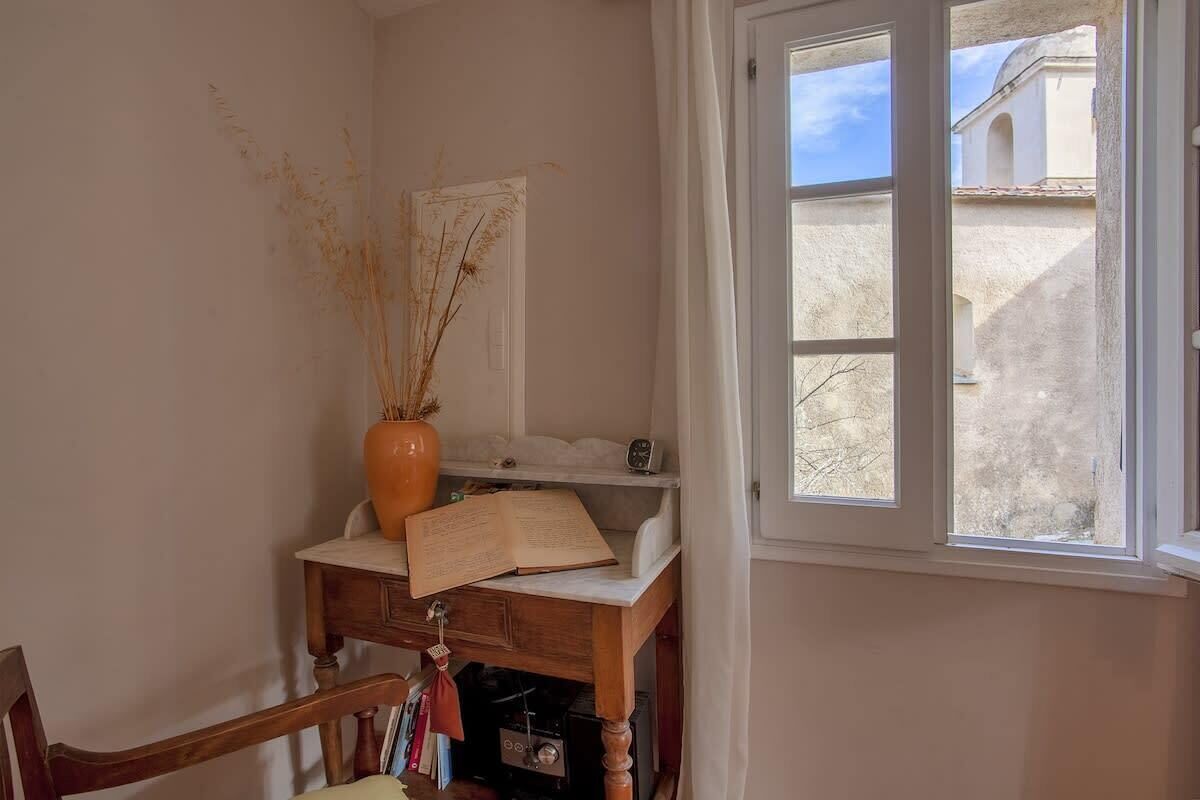 A corner room with a wooden desk, a vase with dried plants, a book, and a window with a view of buildings.