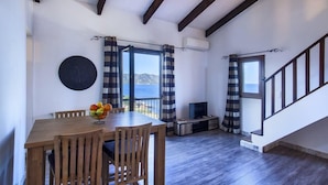 Uma área de refeições com mesa e cadeiras de madeira, uma cesta de frutas e vista para o mar através de grandes janelas.