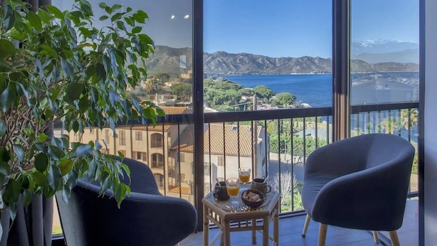 Uma varanda com duas cadeiras, uma mesa pequena com bebidas e uma tigela, e vista para o mar e montanhas.