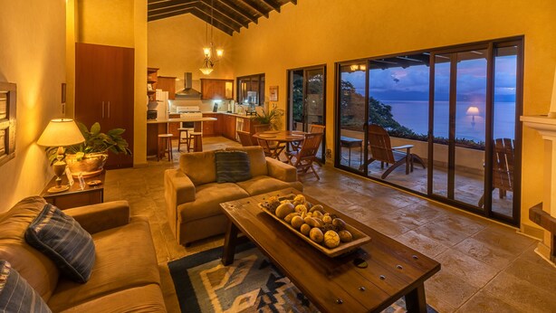 A spacious living area with a large window overlooking a scenic view, a cozy seating area, and a kitchen in the background. - Casa Cholotío lake view, modern, beach access (Panajachel)