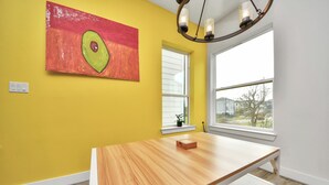 Een kamer met gele muren, een houten tafel, een schilderij van een avocado en een raam met uitzicht.