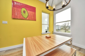 Une pièce aux murs jaunes, avec une table en bois, un tableau représentant un avocat et une fenêtre donnant sur un paysage.