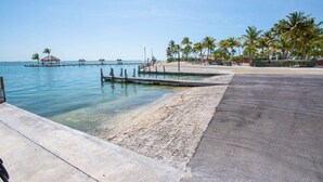 A beach with clear water, a concrete walkway, palm trees, and a pier with huts. - Waterfront Venture Out Home (Cudjoe Key)