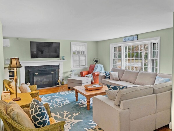 A living room with a fireplace, a sofa, a chair, a table, and a fan. - Family Tides – Your Cape Cod Adventure Base (Centerville)