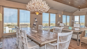 Amplia zona de comedor con una mesa de madera y sillas de mimbre blanco, con vistas a la playa.