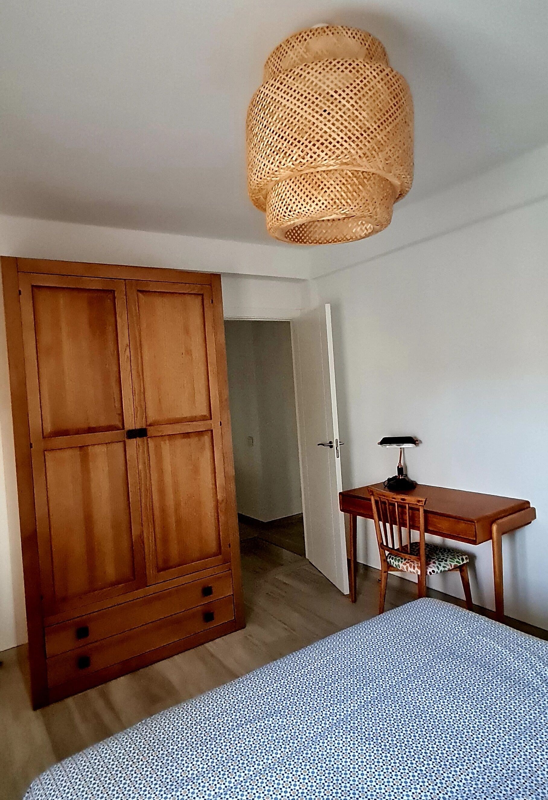 Une chambre avec une armoire en bois, un bureau avec une chaise, une couvre-lit à motifs et un luminaire suspendu au plafond, fait de fibres tressées.
