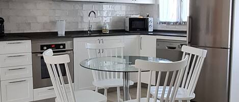 A modern kitchen with white cabinets, a stainless steel refrigerator, a microwave, and a glass dining table with four chairs.