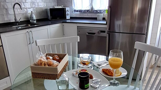 A modern kitchen with white cabinets, a stainless steel refrigerator, a glass dining table with white chairs, and a breakfast setting with bread, juice, and condiments.