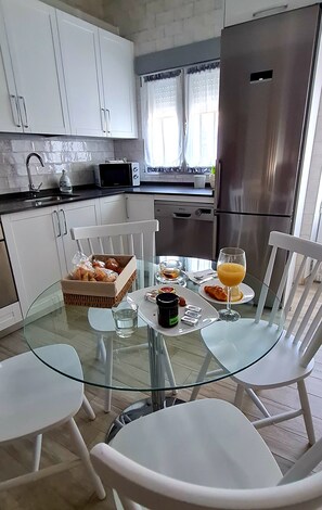 A modern kitchen with white cabinets, a stainless steel refrigerator, a glass dining table with white chairs, and a breakfast setting with bread, juice, and condiments.