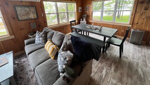 Interior acogedor de una cabaña con techo de madera, un sofá gris con cojines estampados, una mesa de comedor con sillas y ventanas amplias que ofrecen vistas al exterior.