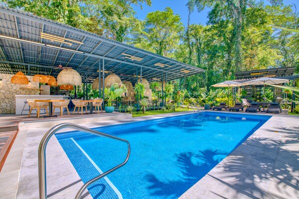 A swimming pool with a metal ladder, surrounded by outdoor seating and umbrellas, with a covered area and lush greenery in the background. - Exōtico Beach Hotel (Cahuita)