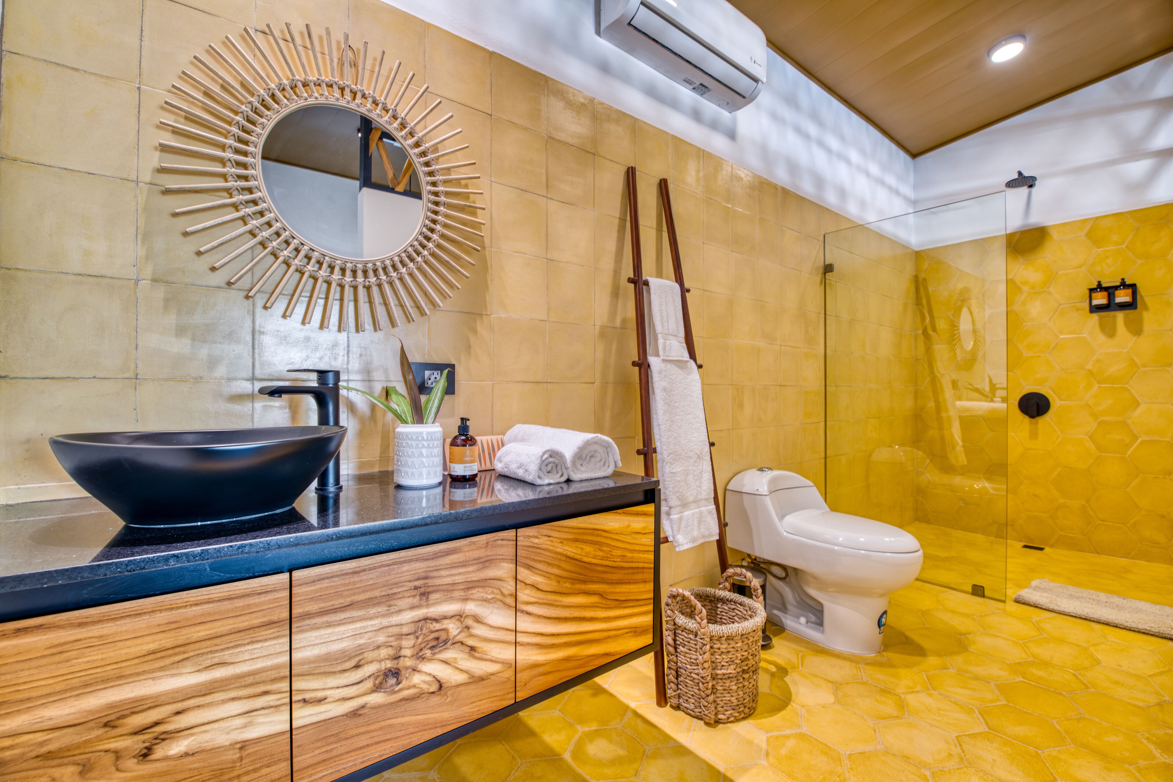 Un baño con tocador de madera, lavamanos negro, espejo, inodoro y zona de ducha.