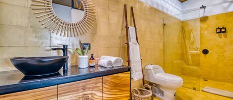 Un baño con tocador de madera, lavamanos negro, espejo, inodoro y zona de ducha.