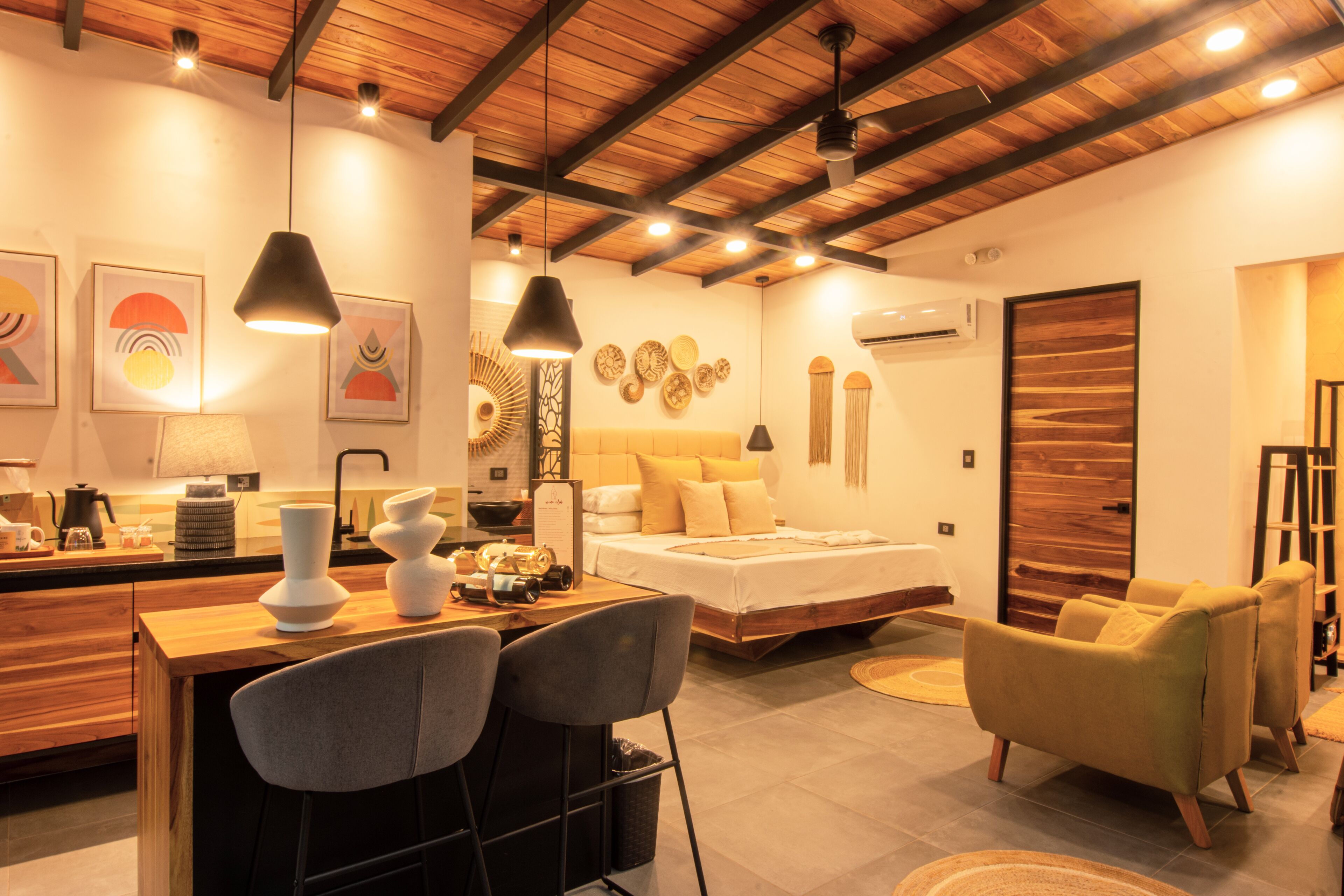 A modern bedroom with a wooden ceiling, a bed with white bedding, a desk with chairs, and artwork on the walls.
