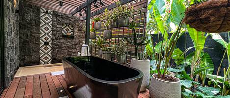 A modern outdoor bathroom with a black bathtub, surrounded by lush tropical plants and a wooden deck.