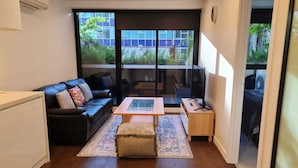 A modern living room with a black leather sofa, a glass coffee table, and a wooden side table. There is a large window providing a view of the outside.