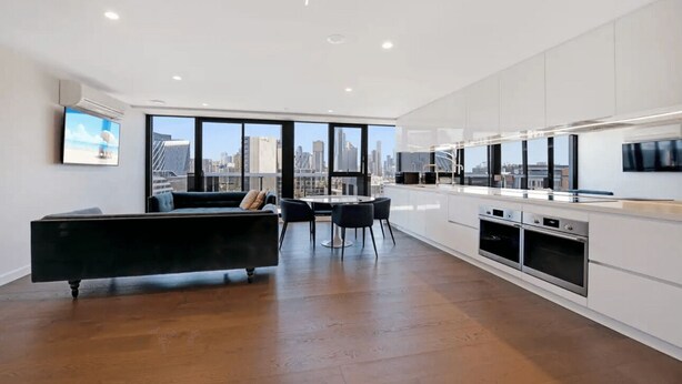 A modern kitchen with white cabinets, a dining area with a black sofa and chairs, and large windows offering a city view.