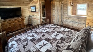 Un dormitorio tipo cabaña de madera con una cama grande, un televisor sobre una cómoda de madera y una ventana con vistas.