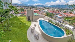 A rooftop pool with a view of a cityscape, surrounded by greenery and a few lounge chairs.