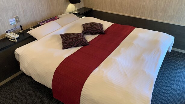 Una cama bien hecha con ropa de cama blanca y roja, dos almohadas estampadas, una lámpara de noche y un teléfono en la mesita.