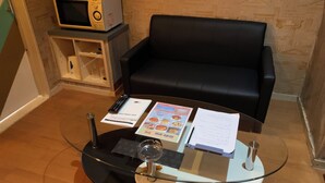 Una mesa con cubierta de vidrio, un control remoto, un menú y unos papeles, un sofá de cuero negro, un microondas y una pequeña estantería.