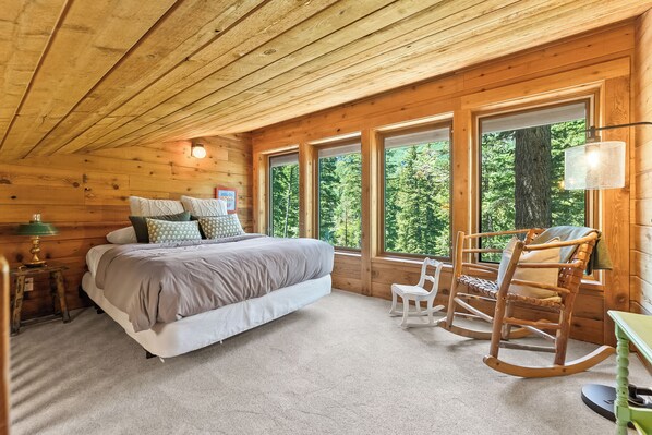 A wooden cabin bedroom with a large bed, a rocking chair, and multiple windows offering a view of the forest. - Carpe Montem - Modern, Views, Large Deck, Hot Tub, Private (Sundance)