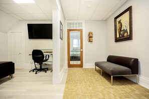 A room with a wooden floor, a grey sofa, a black office chair, a TV mounted on the wall, and a door with a glass panel.