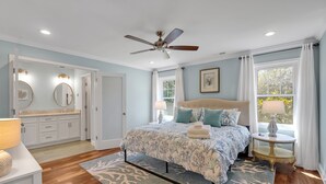 Une chambre à coucher comprenant un lit, un ventilateur de plafond, une table de chevet, une fenêtre avec des rideaux et une salle de bain avec un miroir et un lavabo.