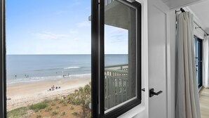 Vista desde una ventana con vista a una playa donde se ven gente y un bote, un balcón con barandilla y una puerta con cortinas.