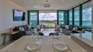 A modern dining area with a view of a golf course, a flat-screen TV on the wall, and a table set for two. - A-402 Stunning one bedroom oceanview (Cupecoy)