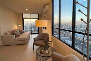 Uma sala de estar moderna com sofá, poltrona e mesa de centro, grandes janelas com vista para a cidade e um lustre.