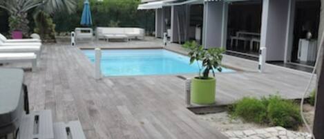 A pool area with a wooden deck, lounge chairs, and a small palm tree.