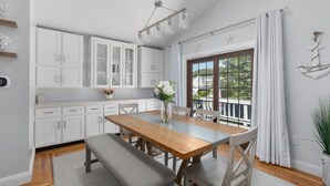 A modern kitchen with white cabinets, a wooden dining table, and a large window with curtains. - Water Views - 3 Min Walk to Private Beach - Luxury 5-Star Rated Beach House (Lower Township)