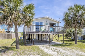 A beachfront house with a wooden deck, a covered porch, and a sandy lawn.