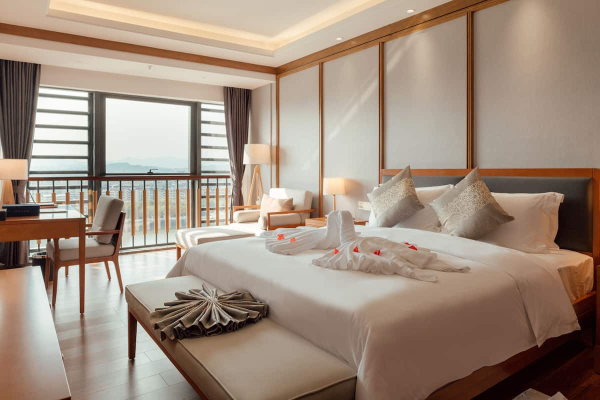 Habitación de hotel con una cama grande, un escritorio, una silla y vistas a la ciudad a través de amplios ventanales.