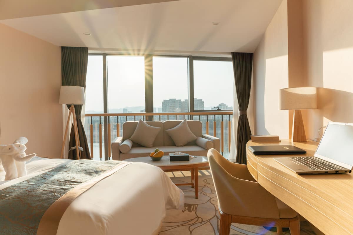 Habitación de hotel con cama, escritorio con una laptop, sofá y vistas de la ciudad a través de amplias ventanas.