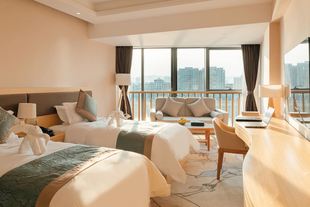 Habitación de hotel con dos camas, un escritorio, una silla y un balcón con vista a la ciudad.