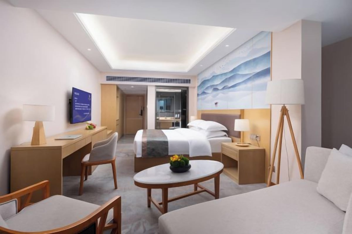 Quarto de hotel moderno com uma cama grande, uma escrivaninha, uma cadeira, uma mesinha e uma televisão.