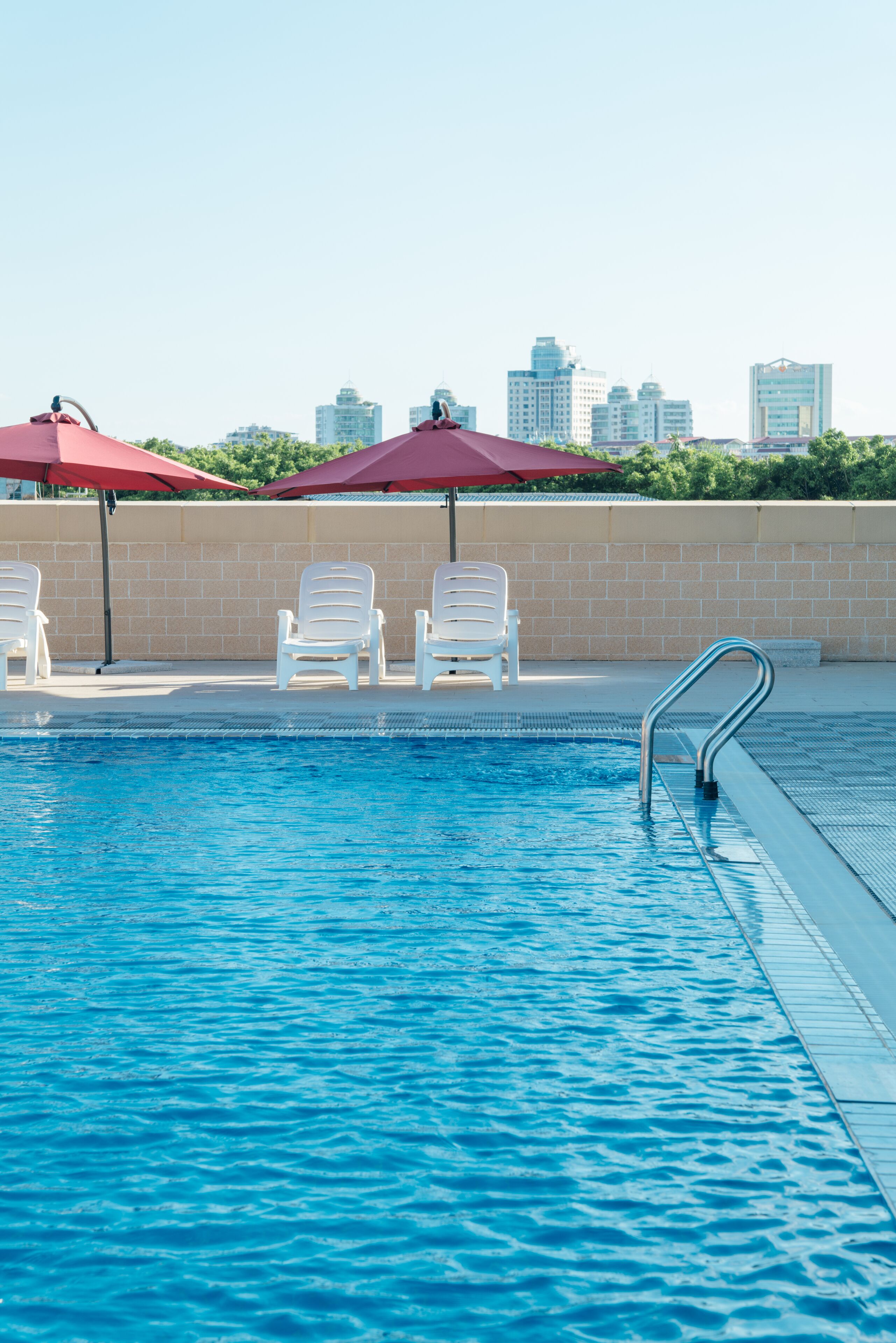 A swimming pool with white lounge chairs and red umbrellas, a metal pool ladder, and a city skyline in the background.