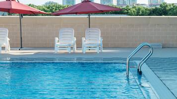 A swimming pool with white lounge chairs and red umbrellas, a metal pool ladder, and a city skyline in the background.